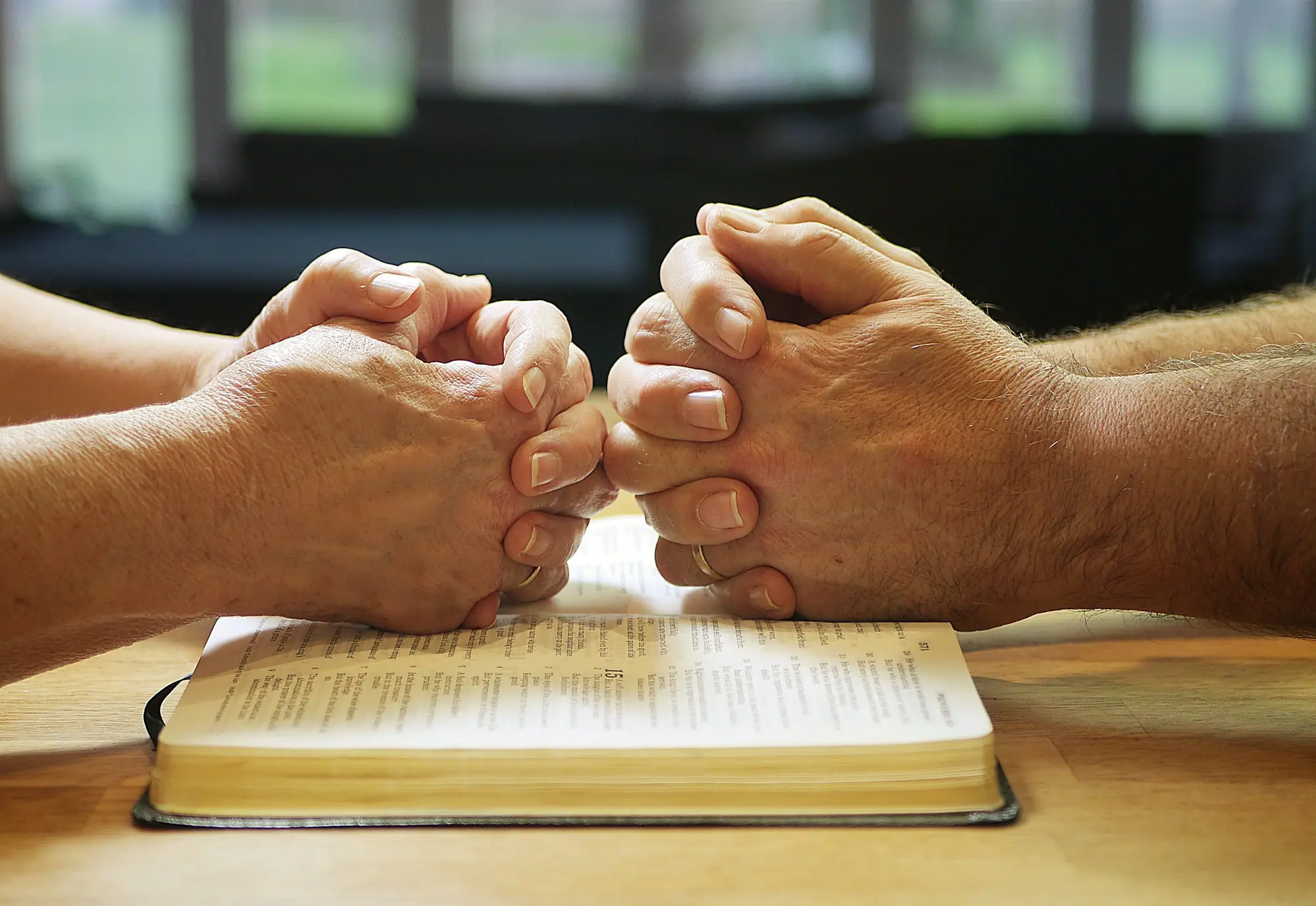 Two people's hands clasped in prayer over an open Bible